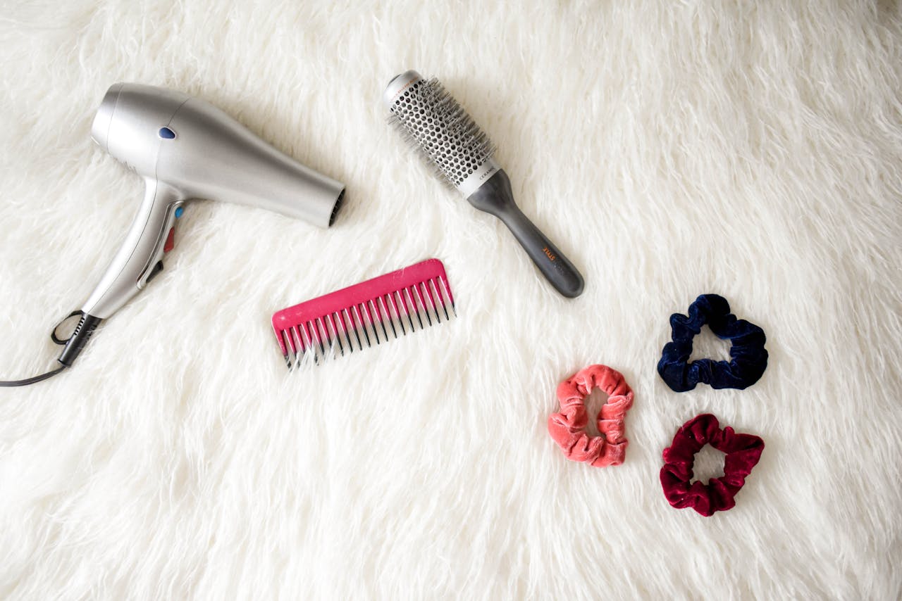 about-01 Flatlay of hairstyling tools including a hairdryer, brush, comb, and scrunchies on white fur.